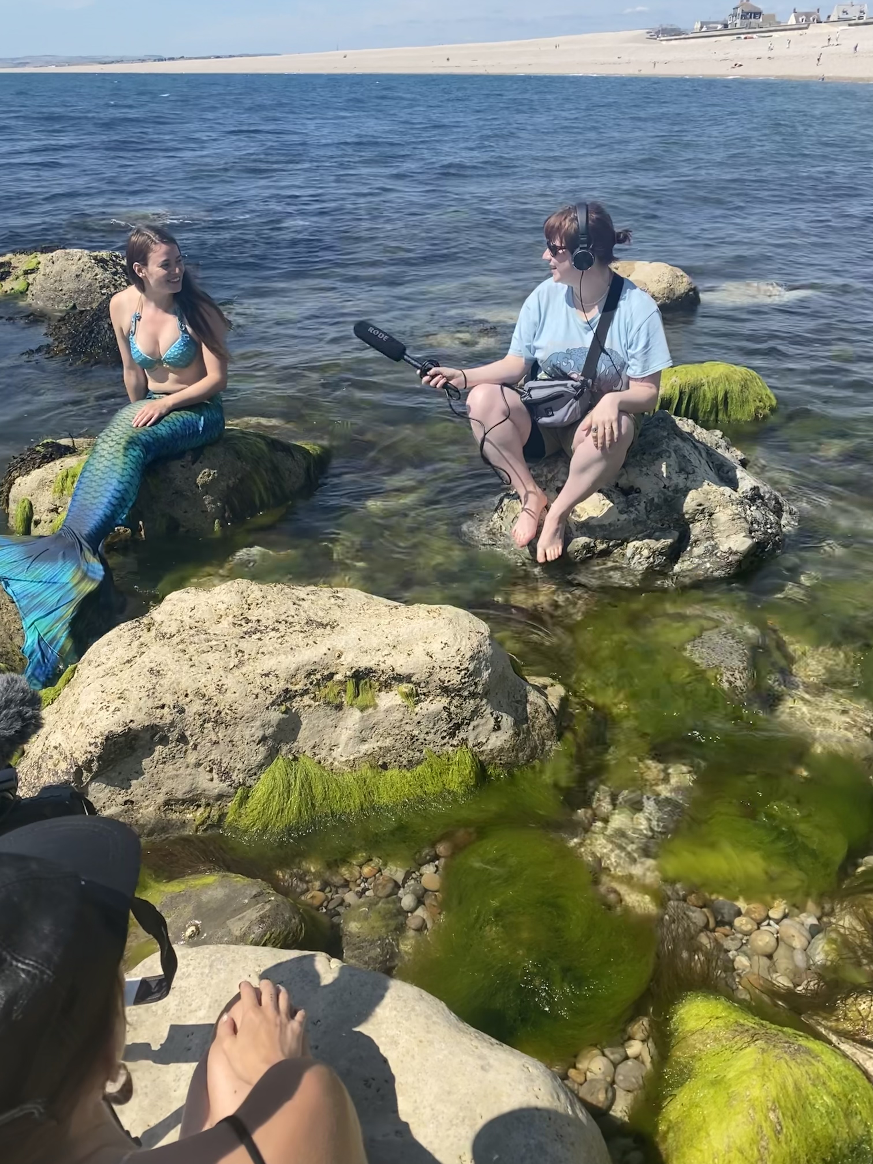Kaia sits on a rock in full mermaid gear during her interview. The sound technician is next to her holding out a boom mic.