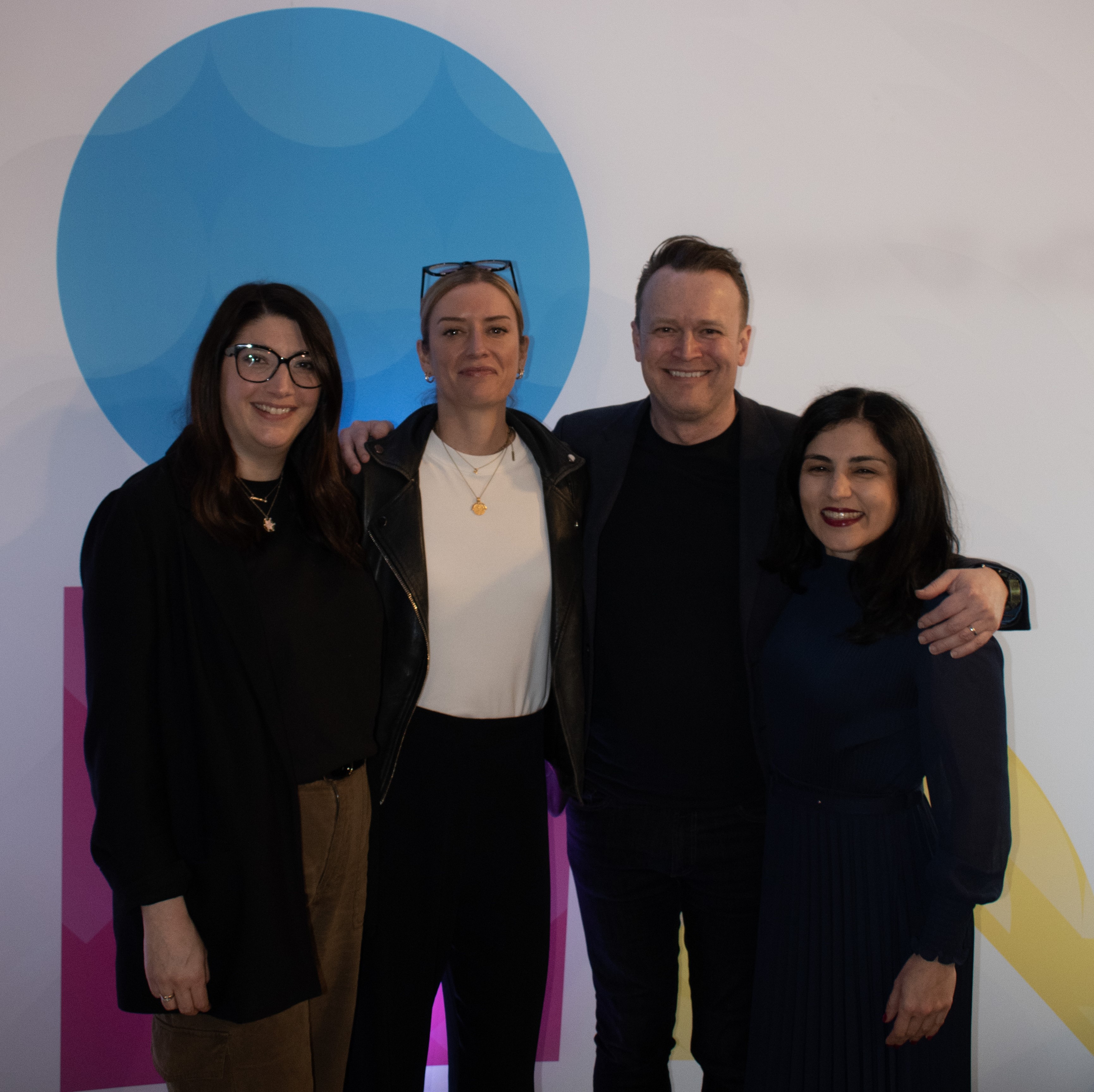 Left to right: Jane, Katie, Paul and Francesca standing with their arms around each other and smiling. Behind them is some blue and pink graphic circles.