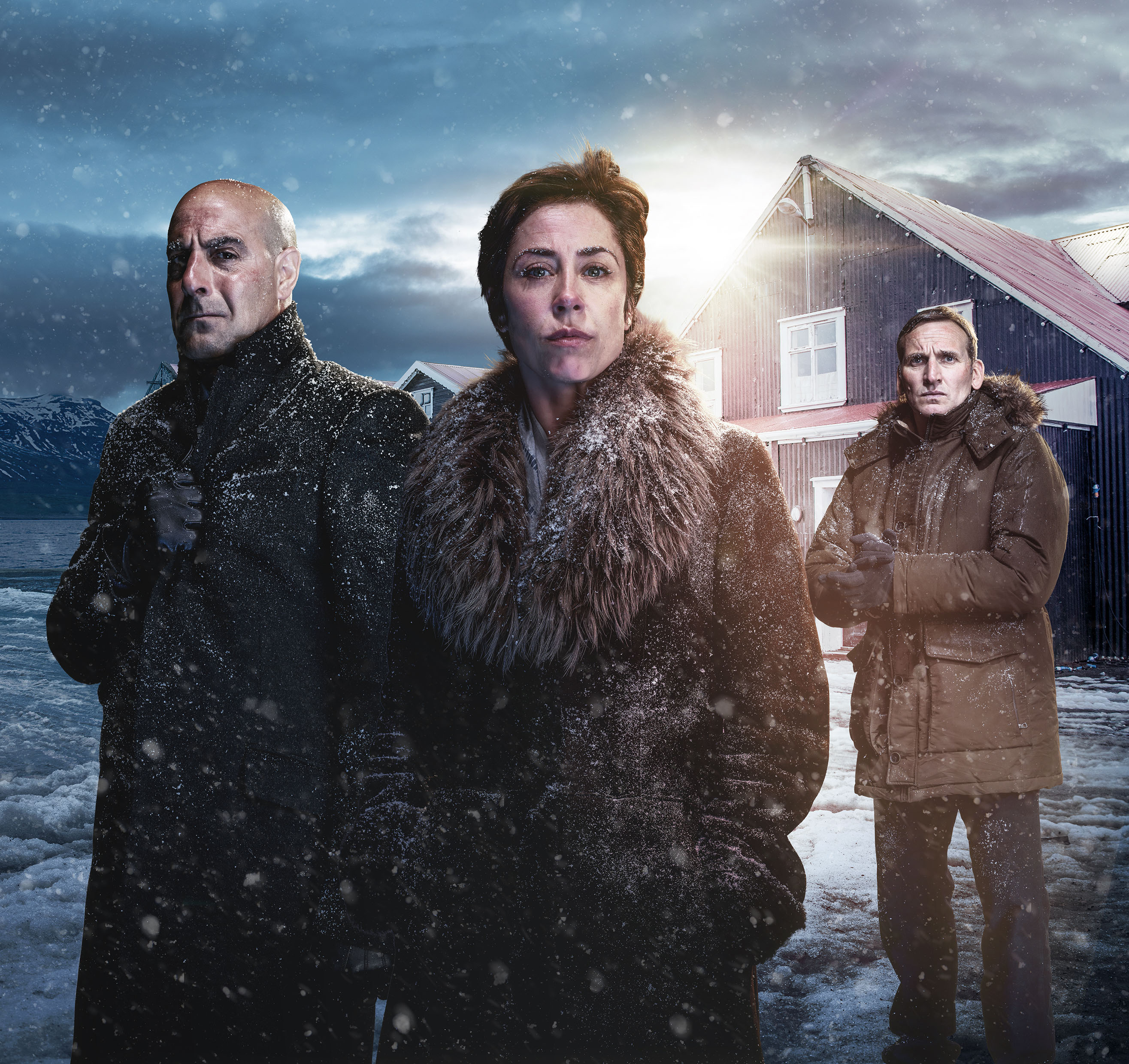 Image courtesy of Sky UK. A posed edited image for Fortitude. The landscape is snowy and dark. Stanley Tucci, a bald tanned man wearing a black scarf and coat stands on the left. In the middle is a white woman with brown hair wearing a fur collar and coat. On the right is a white man with short brown hair wearing a brown jack with a furry collar. Behind them on the right is a timber house with a red roof.