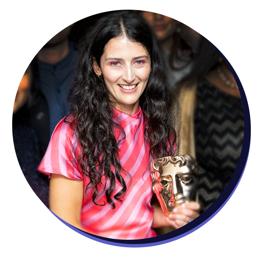 Alex Genova. A white woman with long curly black hair. She is smiling holding a golden bafta award and wearing a stripey pink dress.