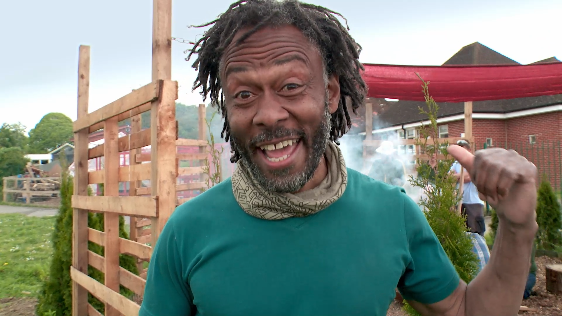 Danny Clarke is smiling open mouth mid-speech and pointing with his thumb behind him. He is wearing a green, turquoise tshirt and a green baclava scarf. He has short greying black locs. Behidn his is a pallet fence and somg lawn. You can see smoke behind him but not the source. There is also a red marquee covering on the right side and houses in the distance.