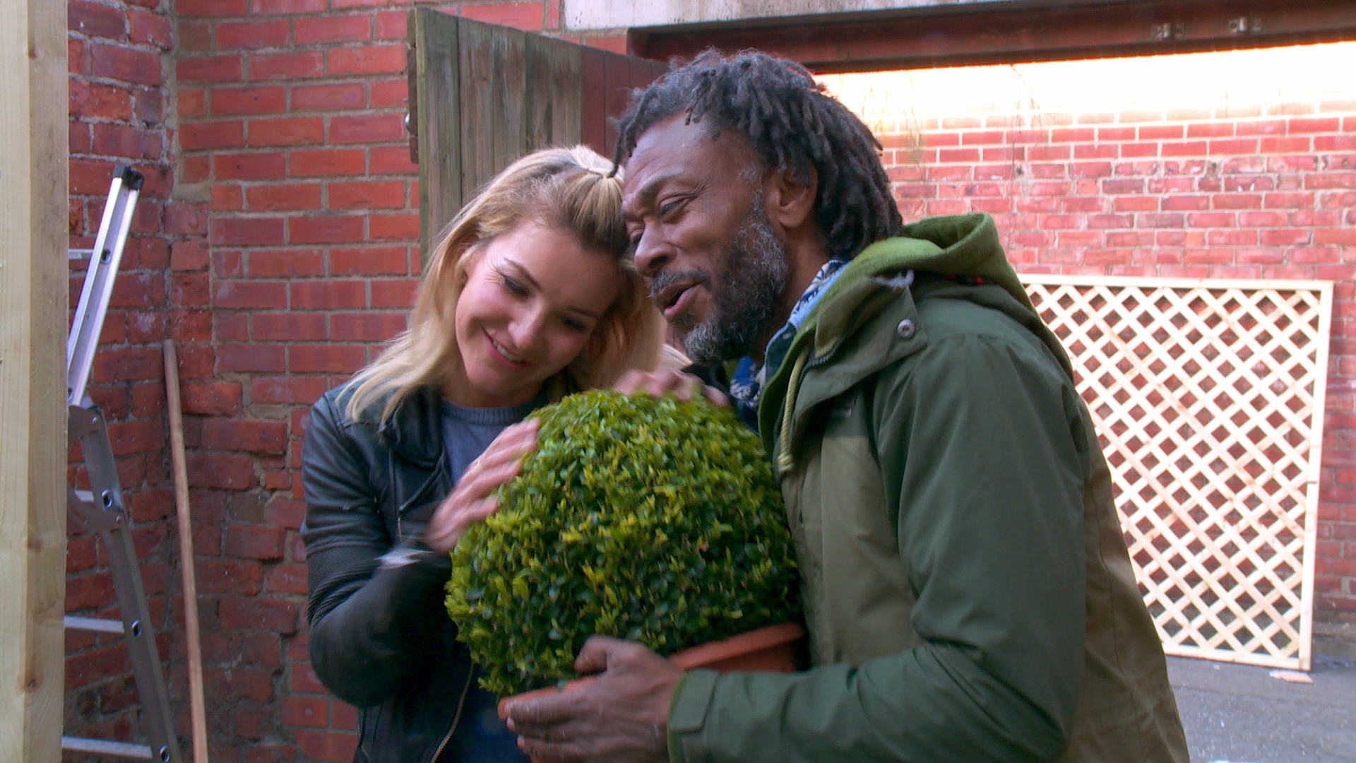 Danny Clarke is holding a round potted shrub and Helen Skelton is petting it and smiliing. Danny is wearing a green mac and has  black greying locs. Helen has blonde shoulder length hair and is weather a leather coat an a blue tshirt. Behidn them is a read bridge wall and a loose lattice fence leaning against the wall.