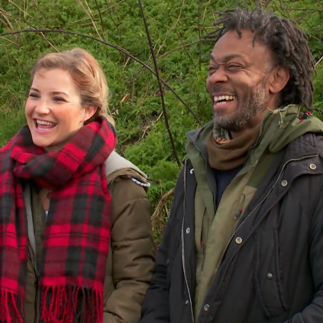 Helen Sketon and Danny Clarke are laughing and smiling towards the left. Helen is wearing her blonde hair back and has a thick red tartan scarf and brown hodoed winter coat. Danny has black greying locs and wearing a green mac with a brown balaclava scarf. Behind them is the greenery  foliage and branches.
