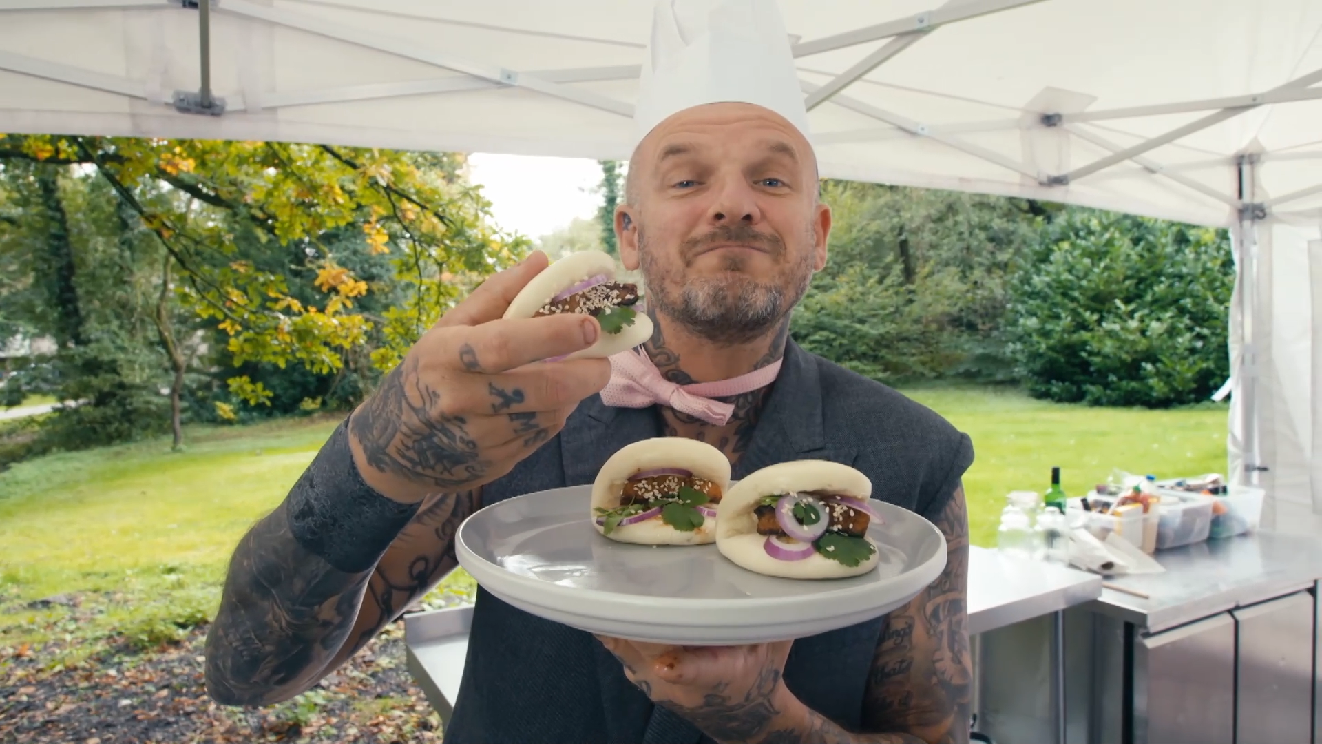 Matt Pritchard wearing a chefs hat in a marque in a garden holding up a bao bun and holding a play with two other bao buns.