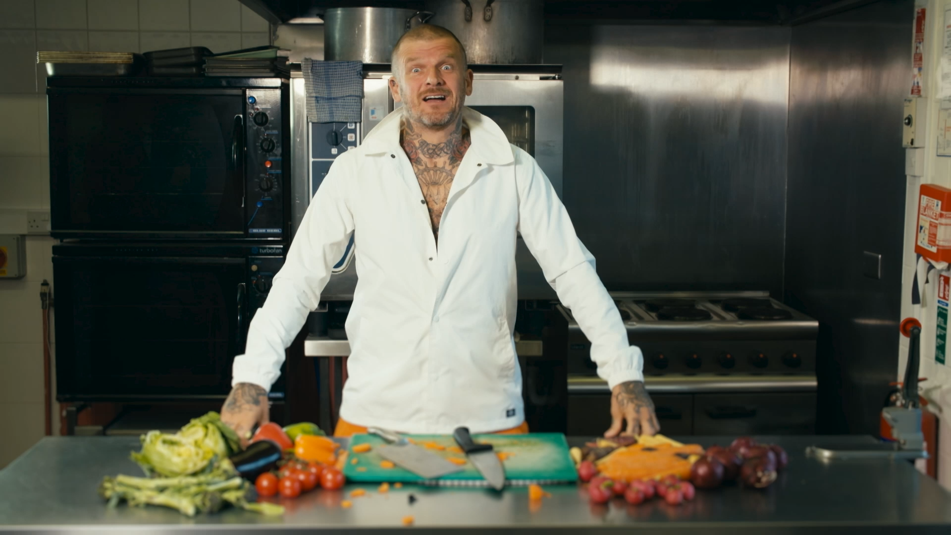 Matt Pritchard in chef whites revealing lots of tattoos standing behind a silver kitchen work bench which is filled with vegetables a chopping board and knife. 