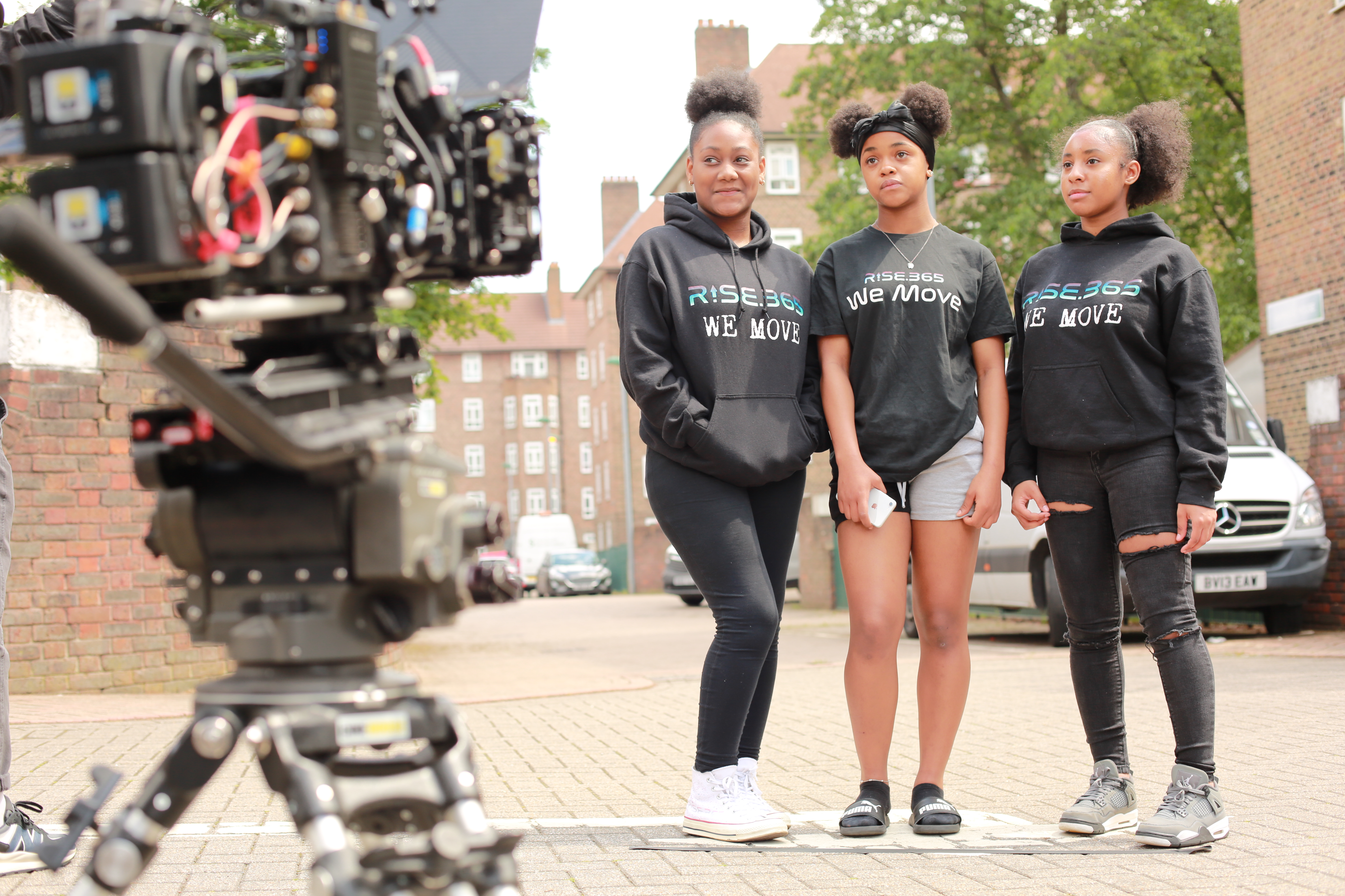On the left is a cfilm camera poitned to three young women standing on the right. These three young women are wearing RISE 365 hoodies and t-shirts, have their afro hair tied up. They are standing in a car park on a London estate.
