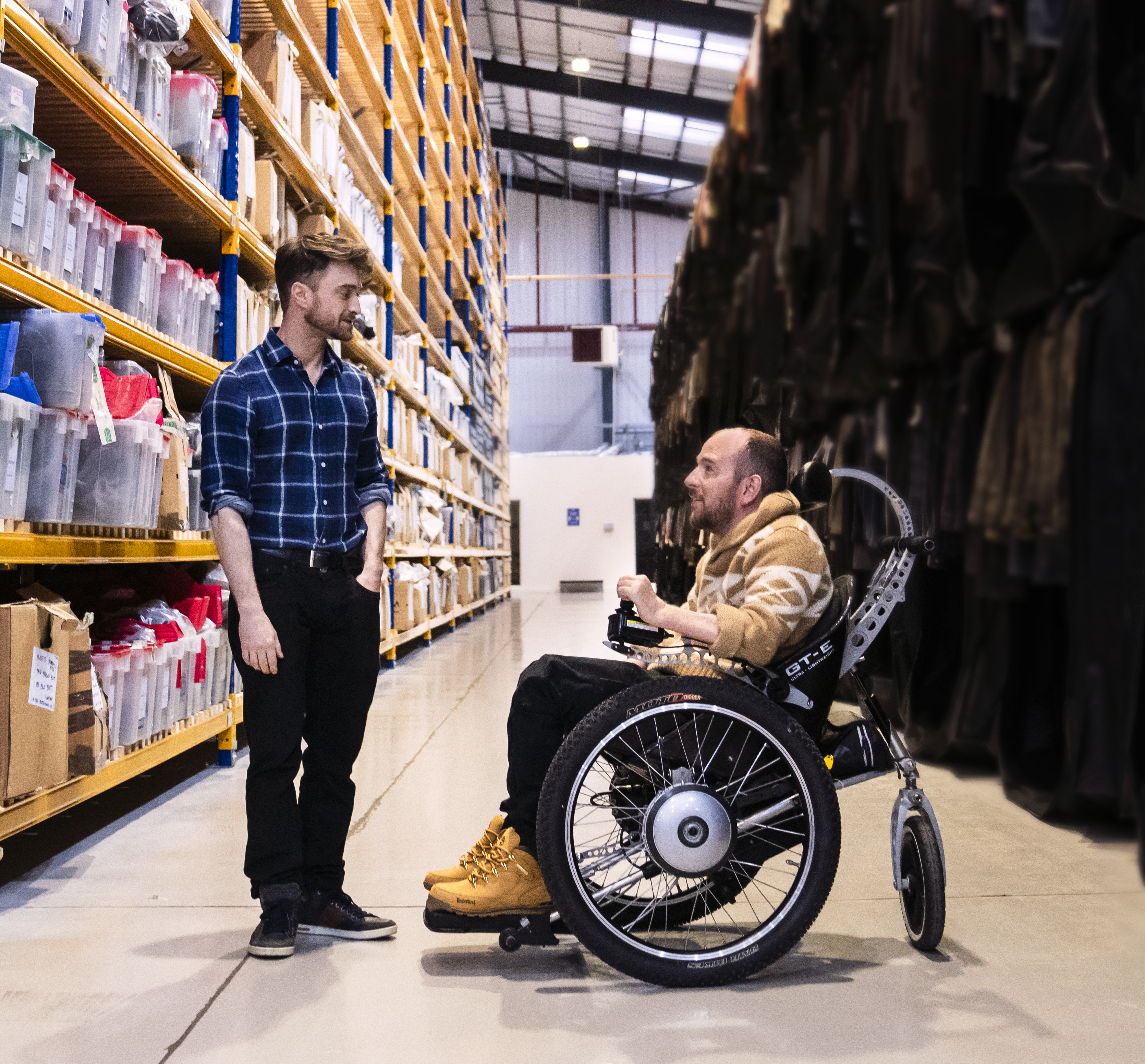 Image courtesy of Sky UK. Image from The Boy Who Lived. Daniel Radcliffe, a brunette white man wearing a blue plaid shirt and black trousers, is standing on the left looking at David Holmes, a balding young man in a wheelchair wearing a yellow hoodie, black jeans and tan boots. They are in a warehouse aisle.