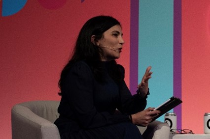 Francesca is mid speech looking to the right sitting in a cream chair. She is wearing a full sleeve navy dress, has black hair and is wearing red lipstick.