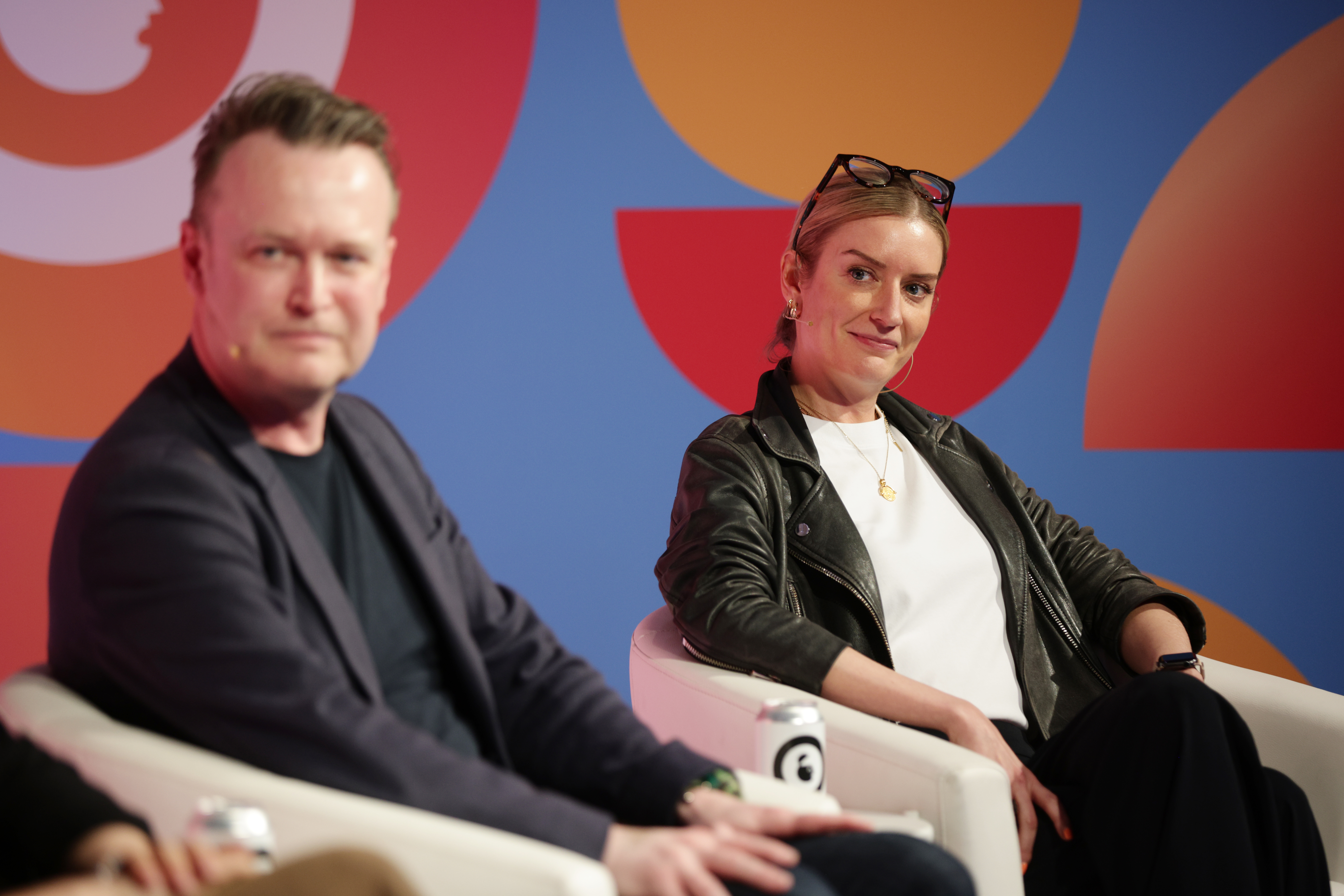 Katie (in focus) and Paul (out of focus) sitting in cream chairs looking to the left and angles to the right. Katie is wearing a black leather jacket and white shirt. She has blonde hair in a pony tail and her black rimmed glasses are on top of her head.