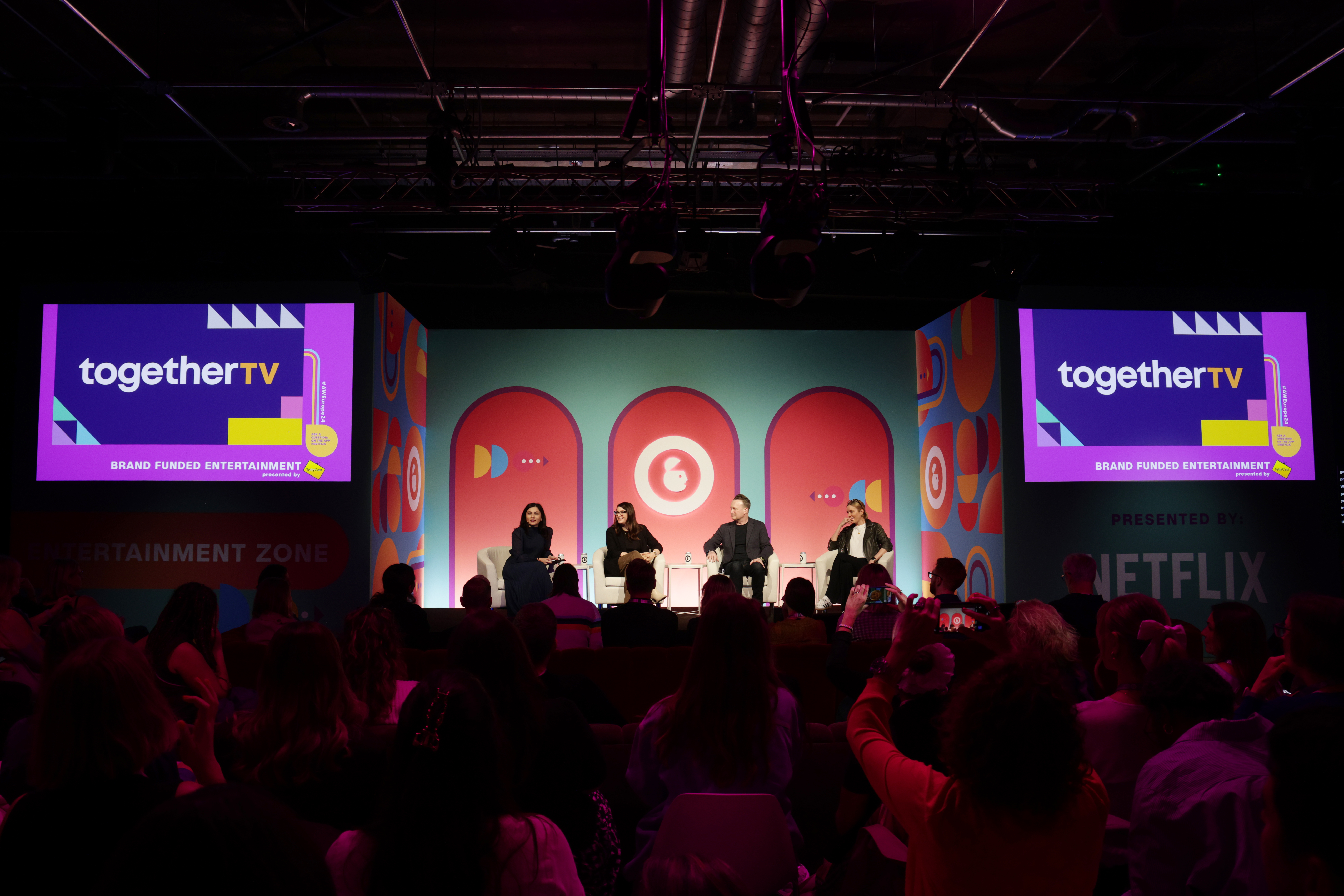 A group of people sitting on a stage with the audience in the foreground and Together TV branding on two screens either side.