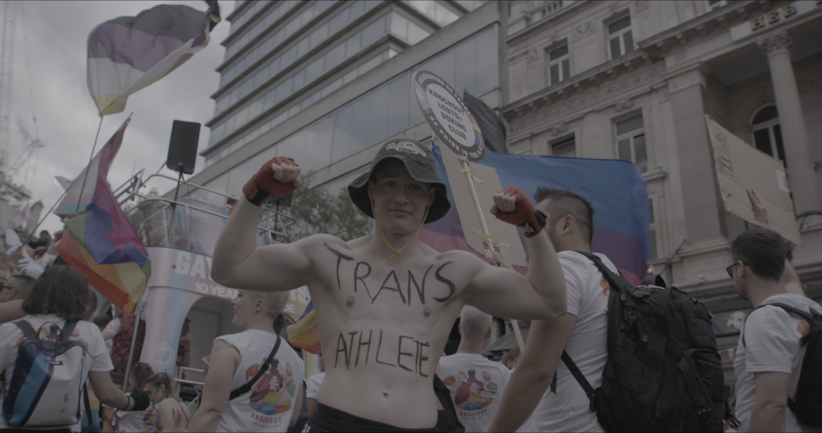 Jill faces the camera at Pride with arms flexed and the words "Trans Athlete" drawn onto his chest.