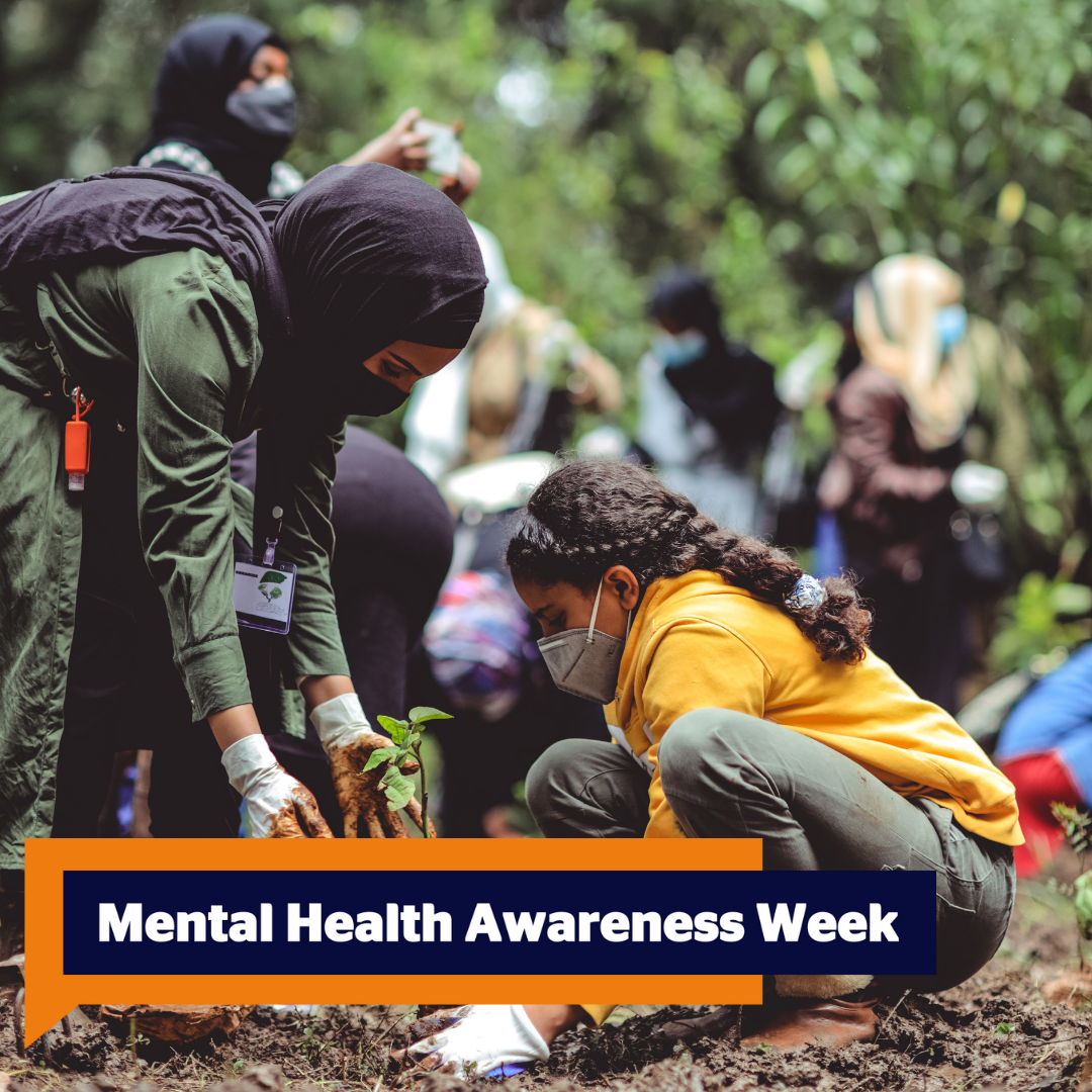 Mental Health Awareness Week text in an orange speech bubble in the bottom left. The photo behind focuses on two women in a crowded community garden planting a young plant into the ground. The woman on the right is crouching, wears her thick grown hair in a plait and a happy orange jumper, green trousers and brown shoes. She is wearing a medical face mask. The woman on the left is bending over to help with a black headscarf and a black surgical face mask. She is wearing a long green shirt dress