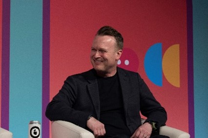 Paul Davies sitting in a cream armchair smiling to the left. He has short cropped brown hair and is wearing a black shirt and blazer.
