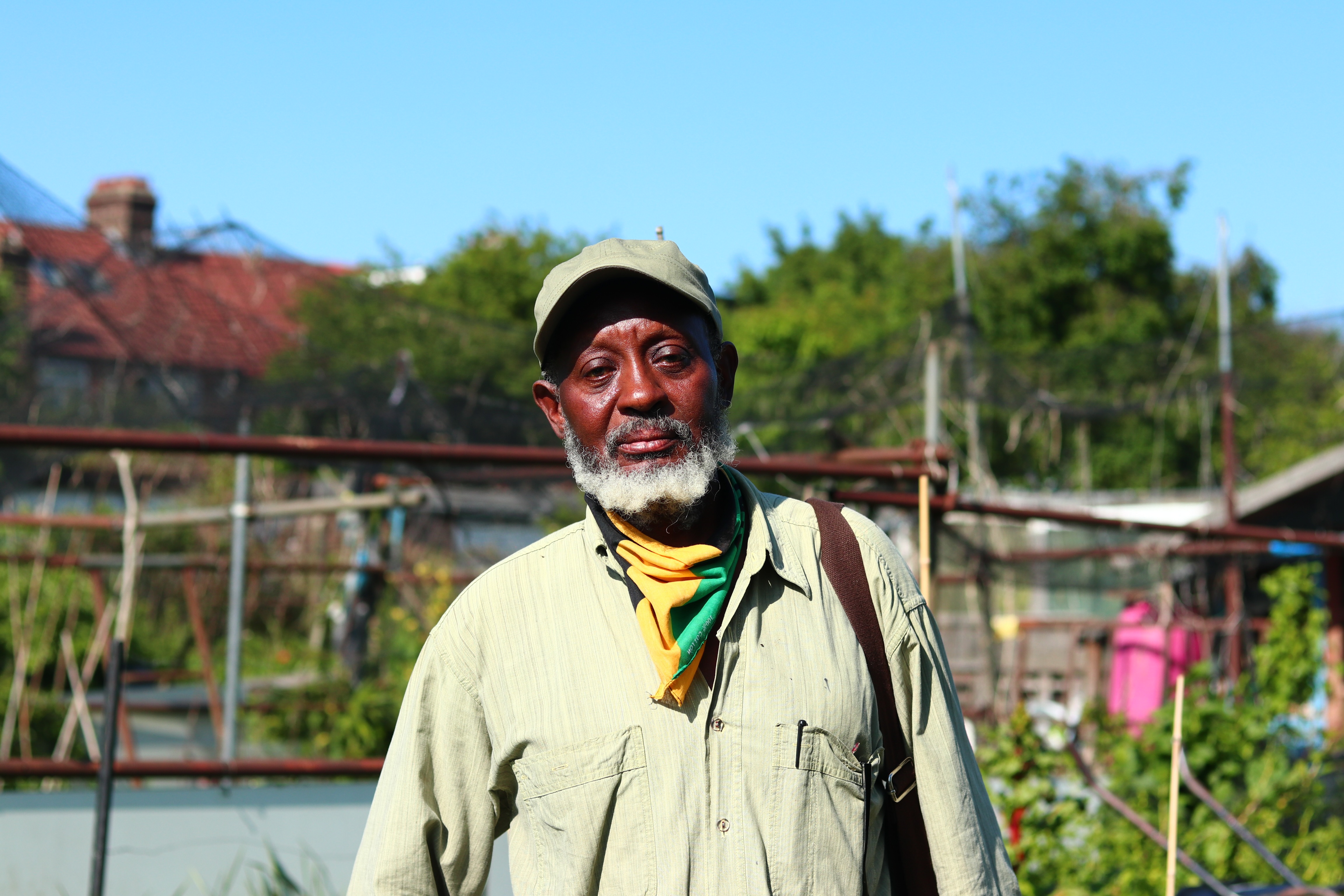 Robbie Samuda is a balck gardener looking at the camera. He is standing in an allotment wearing a khaki cap and shirt and a Jamaica flag balaclava. The sky is blue.