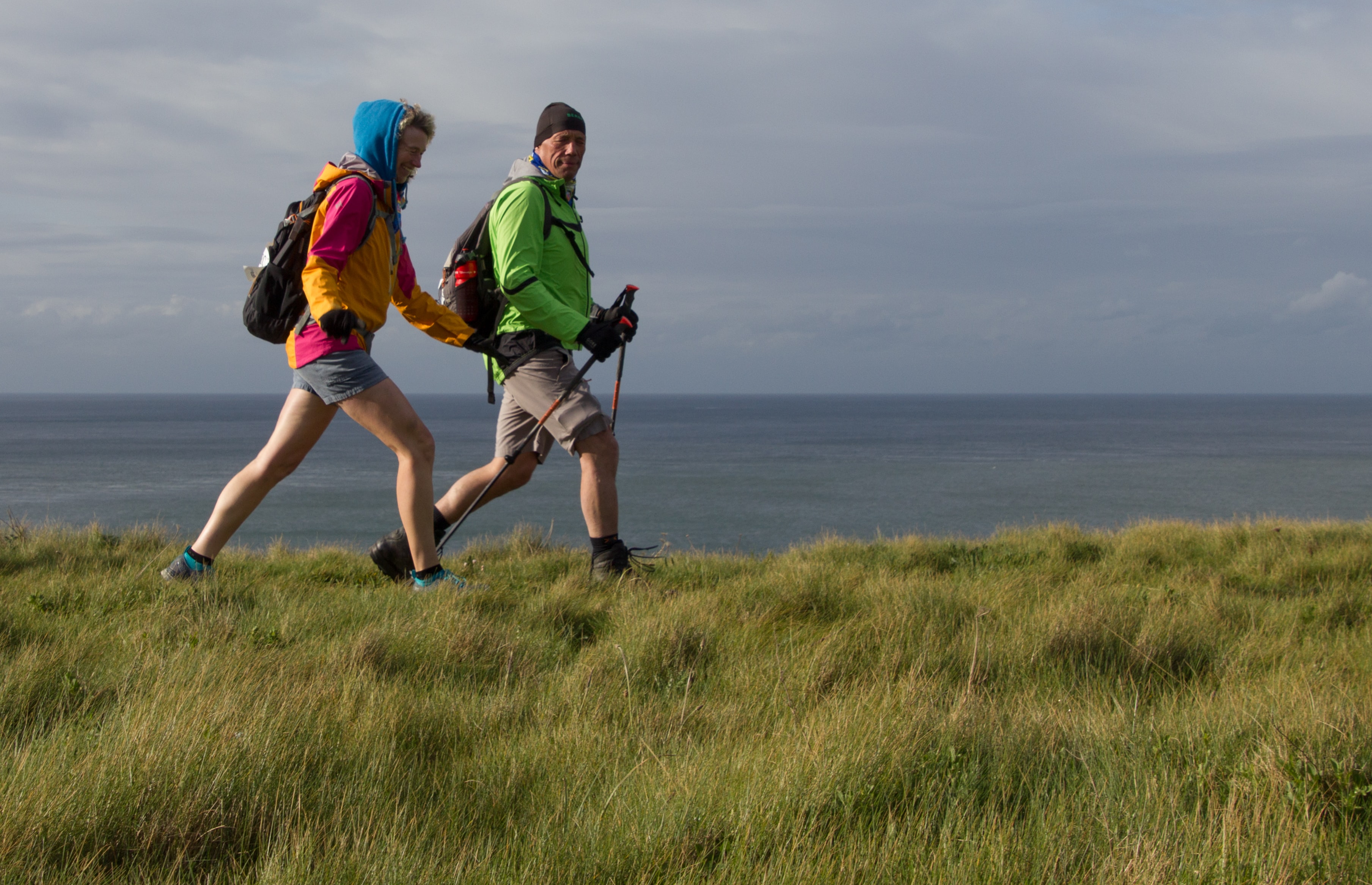 Two people walking in hiking gear along the UK coast. They are walking on grass and in the distance is the sea. The sun is coming from the left. They are wearing shorts and windproof jackets