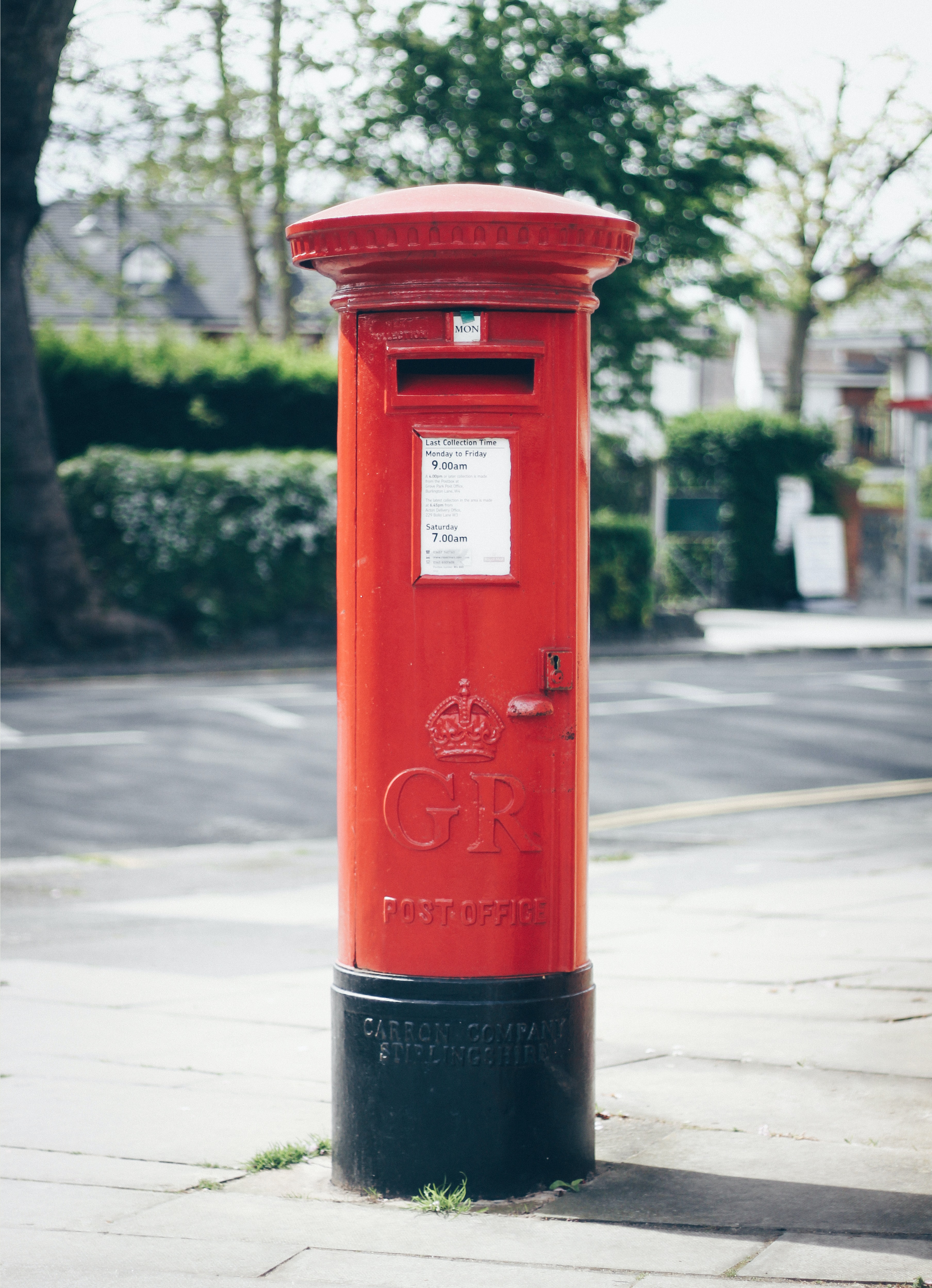 A red UK pillar post box. The background is a blurred street with bushes and trees.