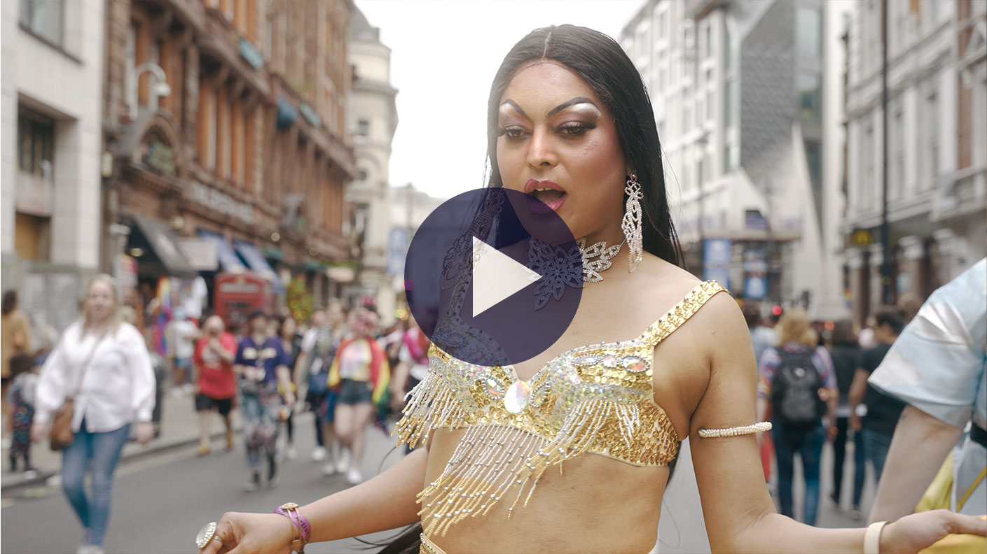 Still from Always, Asifa. Asifa is ddressed in drag with long black wig and a chainmail golden bralette with dangly earrings. She is walking on the street surrounded by people celebrating pride