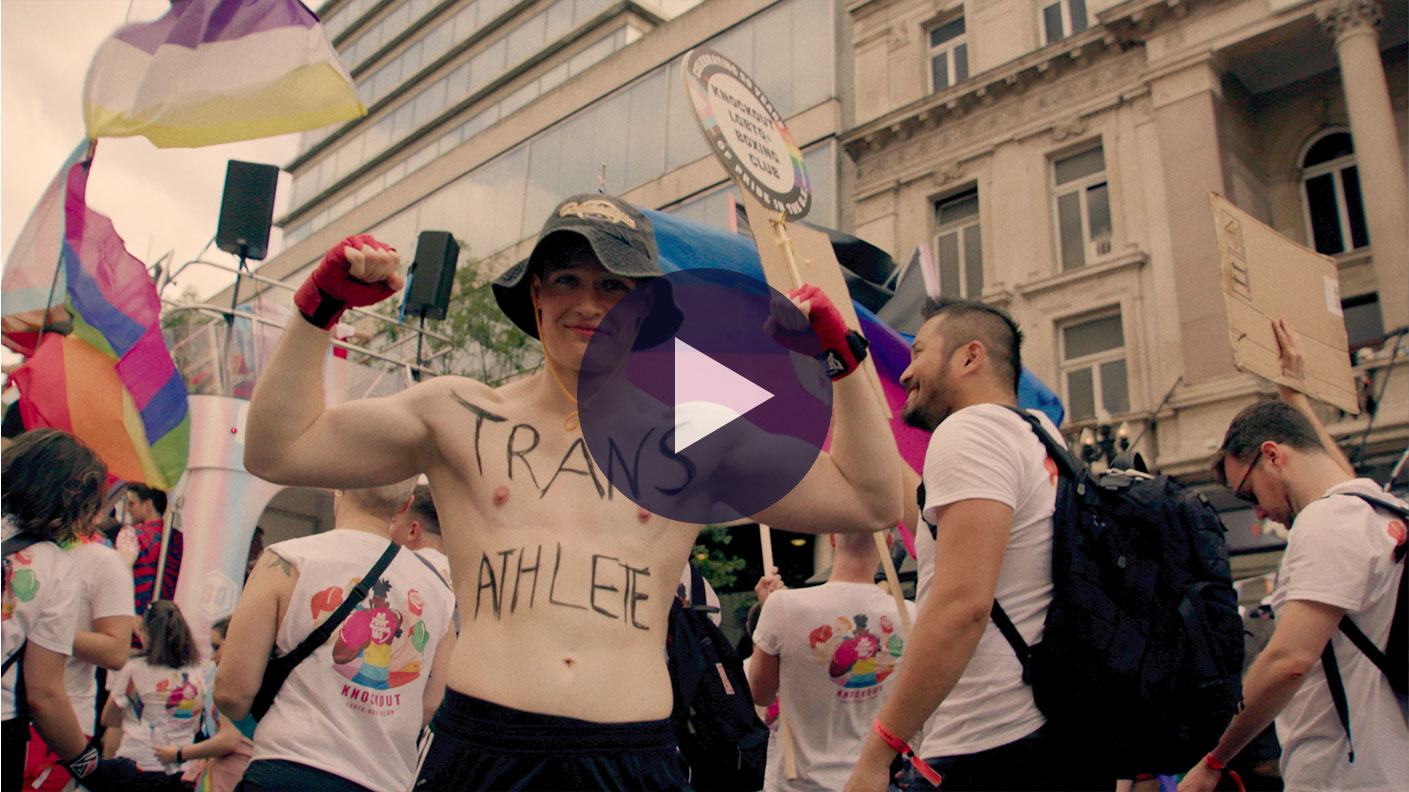 Jill from Come Out Fighting is at a Pride parade. He is smiling at the camera with his arms raised showing of fhis muscles. Written on his chest in black marker is "Trans Athlete". He is wearing a bucket hat and black shorts as well as boxing glvoes. Around him people are walking the opposite direction waving rainbow banners. 